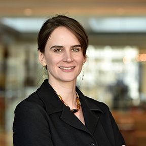 Smiling person in a black blazer standing indoors with a blurred office background