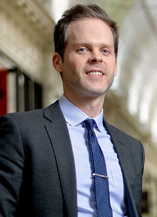 Man in a suit and blue tie standing indoors, smiling at the camera.