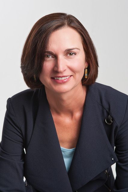 Head-and-shoulders portrait of a smiling woman in a dark blazer against a light gray background