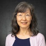 Smiling woman with glasses and shoulder-length dark hair, wearing a navy top and light cardigan on a gray background