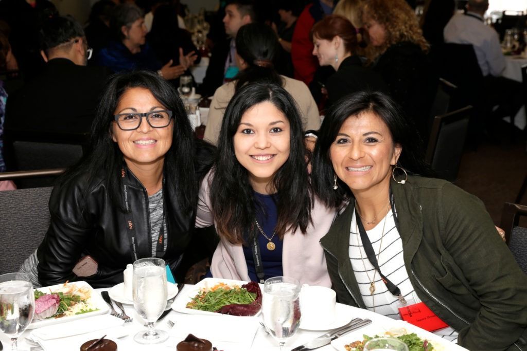 Three people smiling at a dinner table with plates of food and drinks in a dimly lit restaurant.