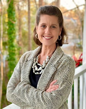 Smiling woman in a patterned jacket stands with arms crossed on a porch