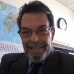 Smiling man wearing glasses and a tie, seated in an office with a world map and clock behind him