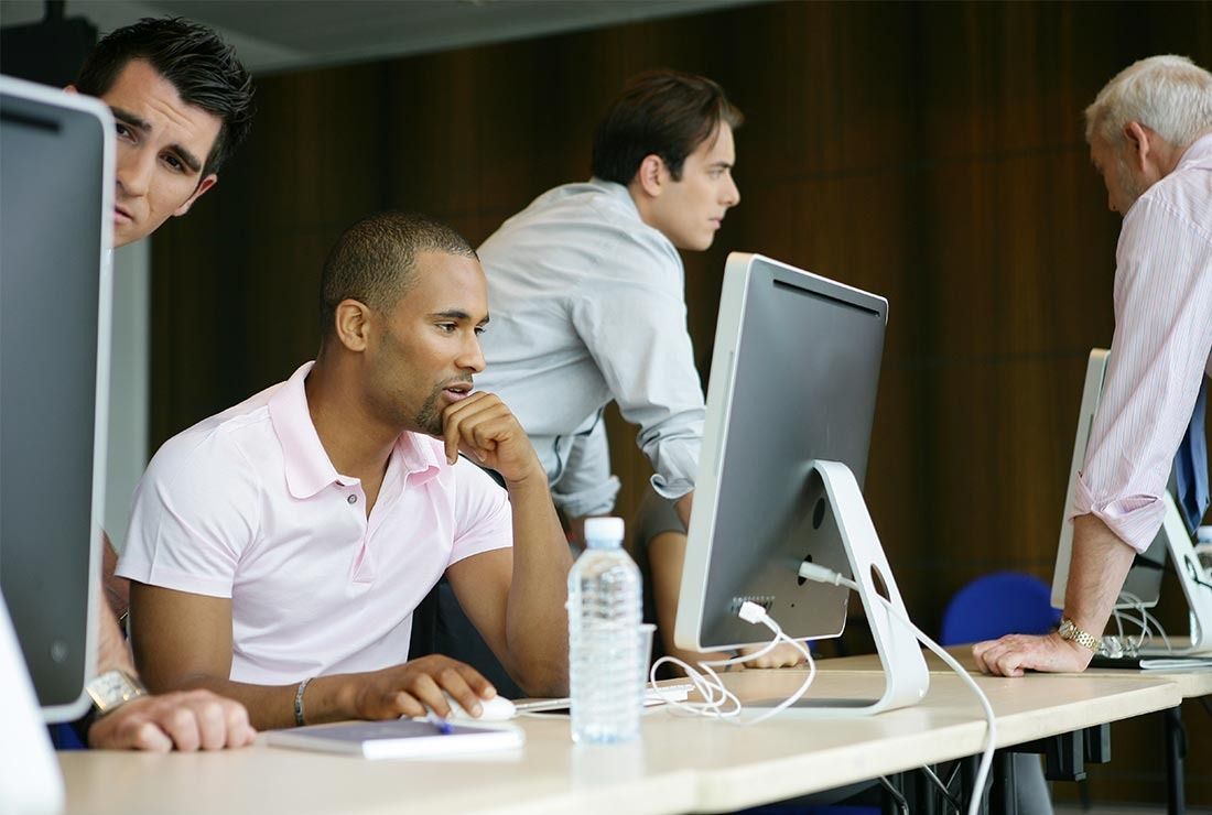 Coworkers in a modern office meeting around desktop computers, one man seated in the foreground.