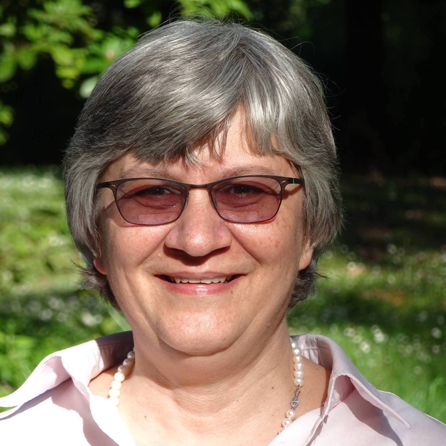 Smiling person wearing glasses outdoors in sunlight, with greenery in the background