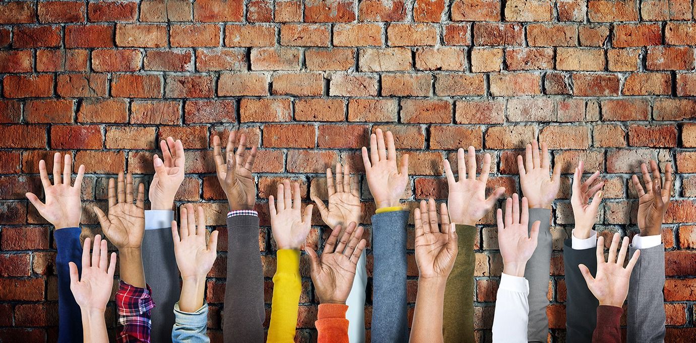 Raised hands of a diverse group in colorful sleeves against a brick wall