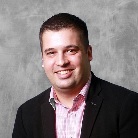 Smiling man in a black blazer and pink striped shirt against a gray studio backdrop