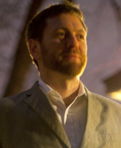 Man in a light suit and white shirt, standing outdoors at night with a thoughtful expression.