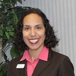 Smiling professional in brown blazer and pink shirt with name tag, standing indoors near a plant.
