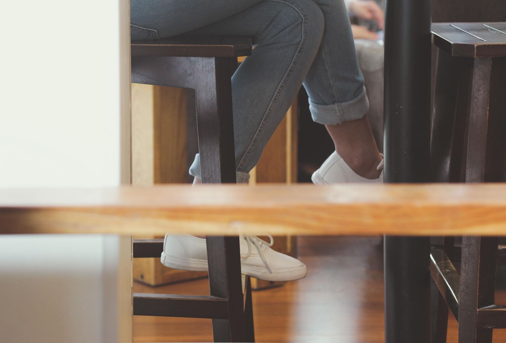 Person’s legs under a table, wearing jeans and white sneakers in a café.