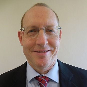 Smiling man in glasses, suit jacket, shirt, and red tie against a plain wall