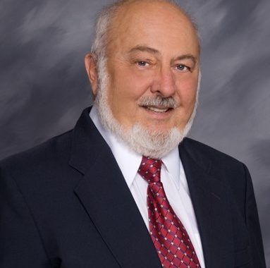Studio portrait of a man in a dark suit, white shirt, and red tie, smiling against a gray backdrop
