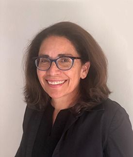 Smiling person with shoulder-length dark hair and glasses, wearing a black top against a light wall.