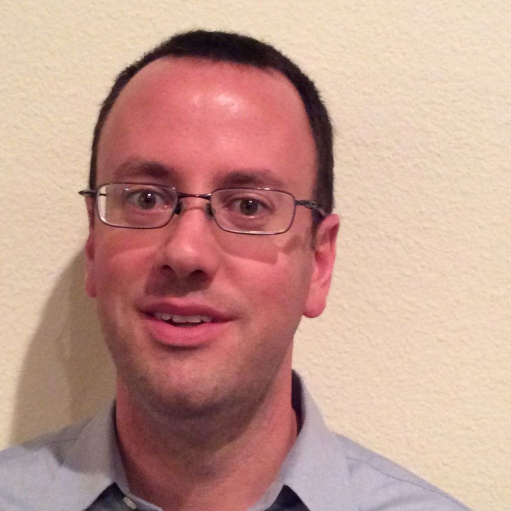 Man wearing glasses and a light blue shirt, smiling against a beige wall.