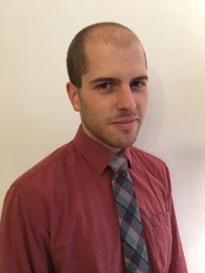 Man in a red shirt and plaid tie standing against a plain wall.