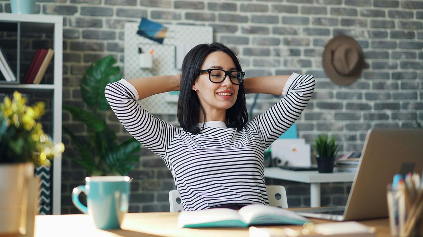 Smiling woman in striped shirt relaxing at a desk with laptop, notebook, and mug in a cozy home office