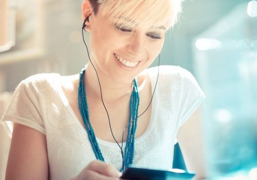 Smiling person with short blonde hair and blue necklace using earphones to look at a phone outdoors