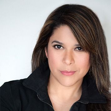 Portrait of a person with shoulder-length dark hair, wearing a black jacket against a light background