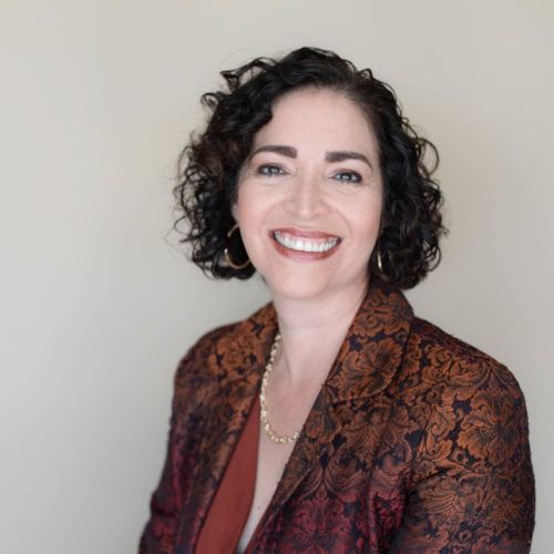 Smiling woman with short curly dark hair in a patterned jacket against a neutral background