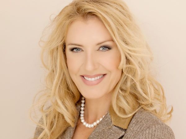 Smiling blonde woman with wavy hair, wearing a pearl necklace and tan jacket on a light background