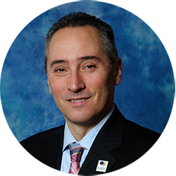 Professional headshot of a man in a dark suit and striped tie against a blue backdrop