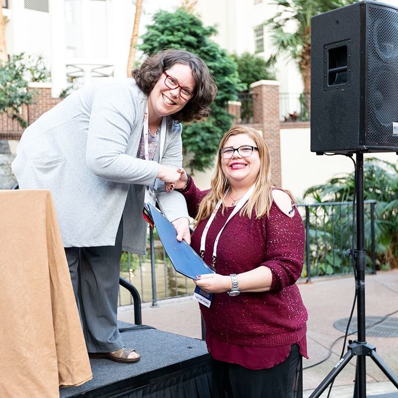 Two people onstage smiling while signing a document at an outdoor event