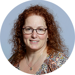 Portrait of a smiling woman with curly red hair, glasses, and a patterned blouse on a blue background
