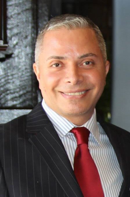 Smiling person in a dark pinstripe suit with a red tie against a dark background