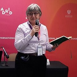 Speaker holding a microphone and booklet at a conference, standing before a red backdrop