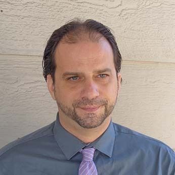 Man in a blue shirt and purple tie posing against a beige wall, with a slight smile