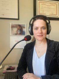 Woman wearing headphones speaking into a microphone in an office, with framed certificates on the wall.