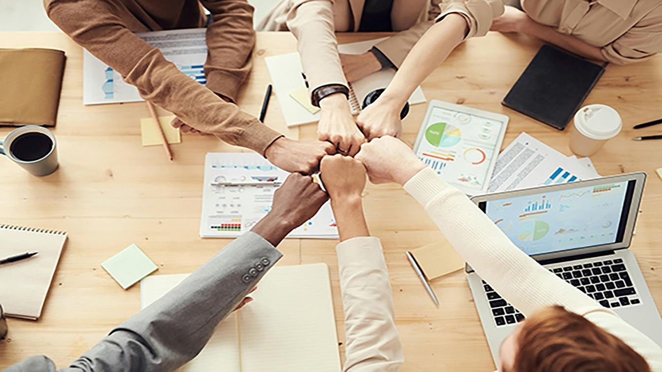 Hands stacked in a team huddle over a conference table with charts, laptop, and coffee cups