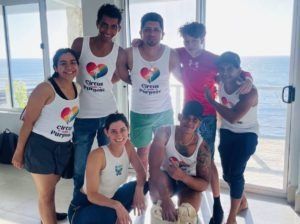 Group of seven people posing indoors by a beach window, wearing matching white tops with a colorful heart logo.