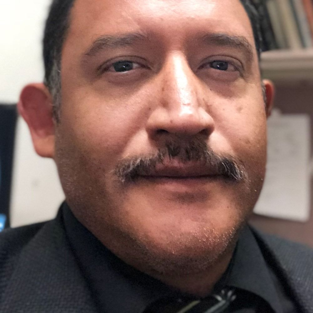 Close-up portrait of a man with a mustache, wearing a dark shirt and tie, against a blurred indoor background