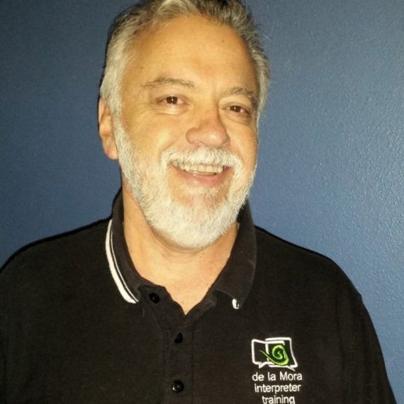 Smiling man with gray hair and beard wearing a black polo shirt against a blue background