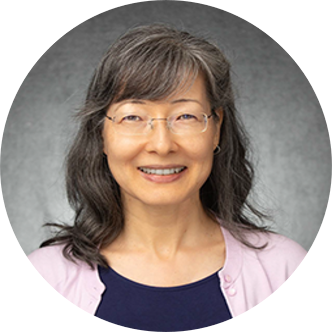 Smiling person with glasses and shoulder-length dark hair against a gray backdrop