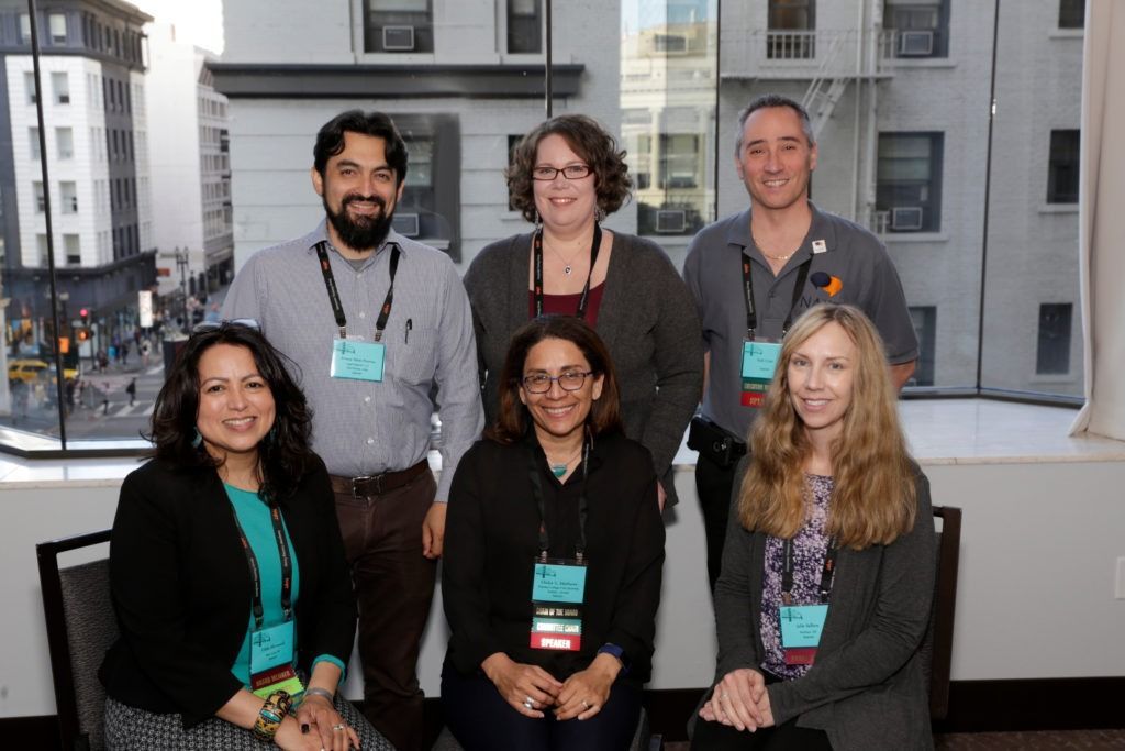 Seven people wearing conference badges pose indoors in front of a city view window.