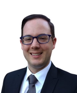 Smiling person in a dark suit, light shirt, tie, and blue glasses on a white background