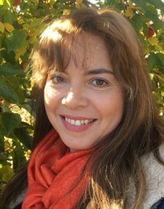 Portrait of a woman smiling, wearing an orange scarf, with green leaves in the background.