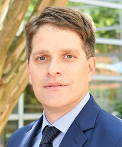 Portrait of a man in a blue suit and tie, standing outdoors with a blurred glass-and-tree background.