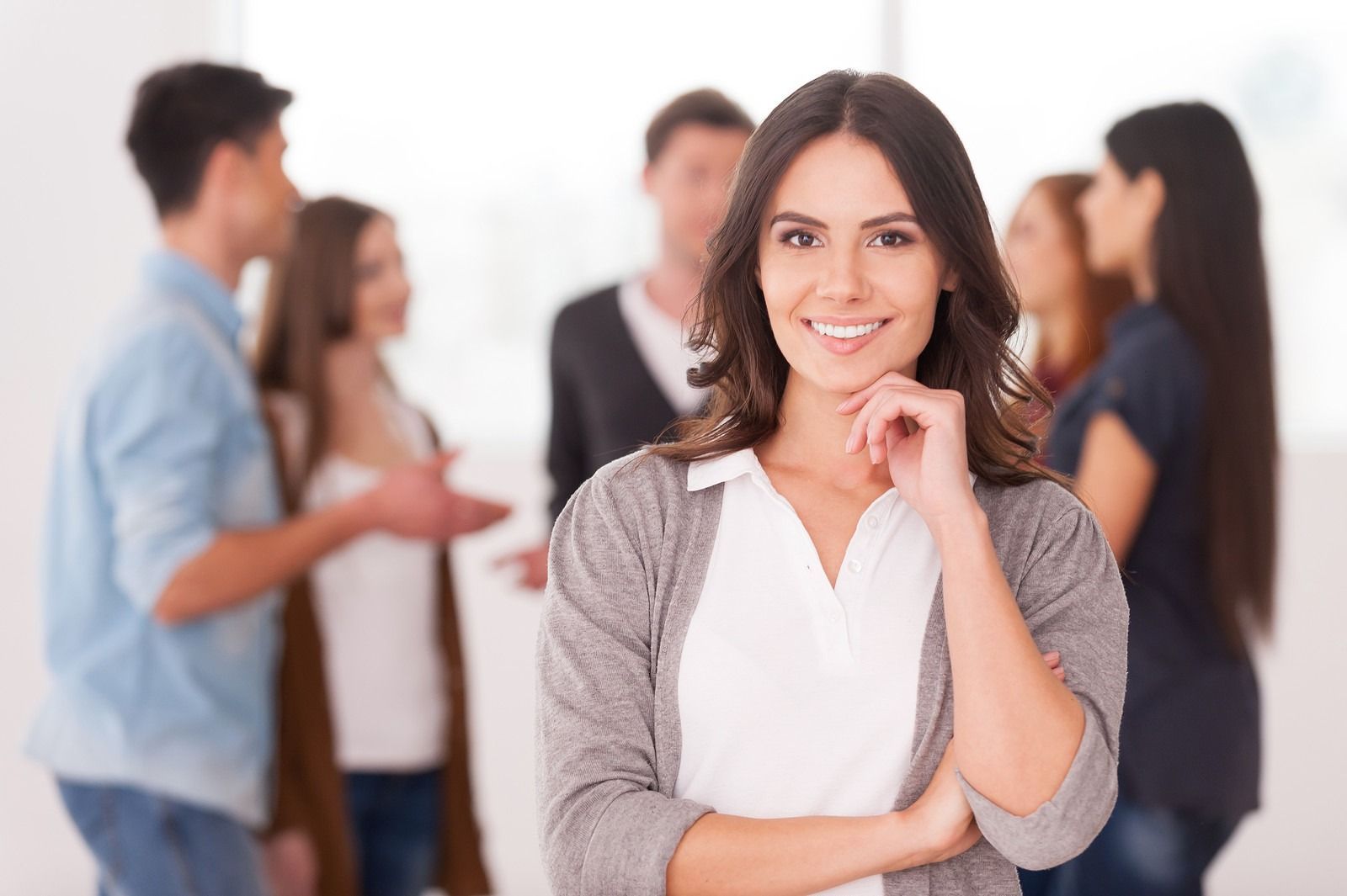 Smiling woman in a white shirt and gray cardigan stands with coworkers in a blurred office setting