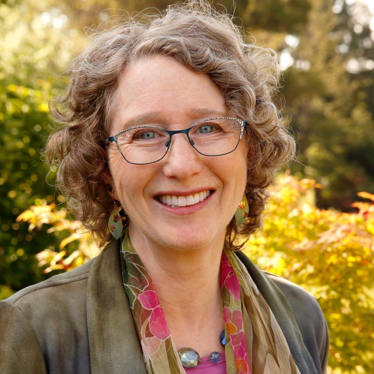 Smiling person with glasses and curly hair outdoors in a garden, wearing a green jacket and patterned scarf