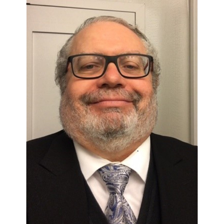 Smiling man in glasses and suit with patterned tie against a plain indoor background