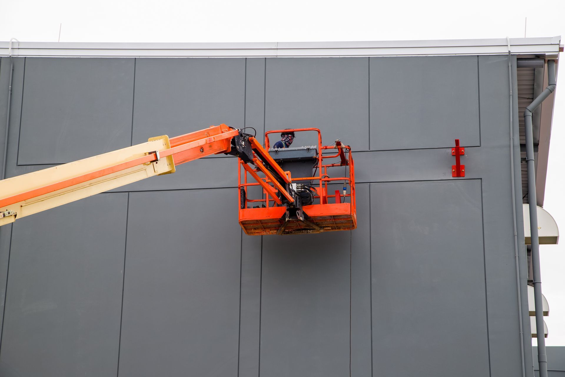 Un ouvrier, dans une nacelle orange, inspecte le mur gris d'un bâtiment près de la ligne de toit.
