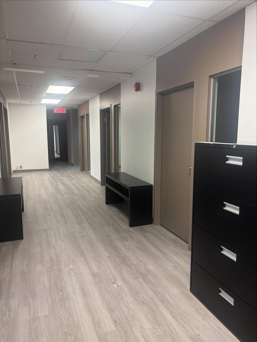 Couloir de bureau vide avec sol gris, portes beiges, armoires noires et éclairage fluorescent au plafond.