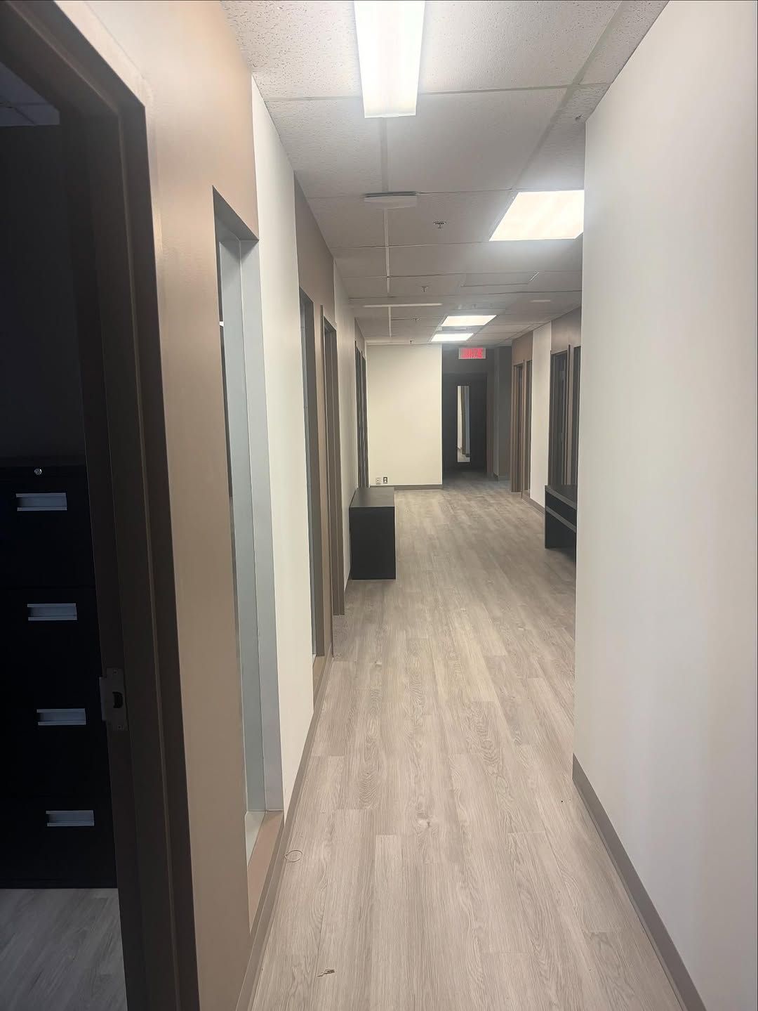 Couloir de bureau avec murs blancs, parquet et éclairage au plafond menant à une porte au fond.