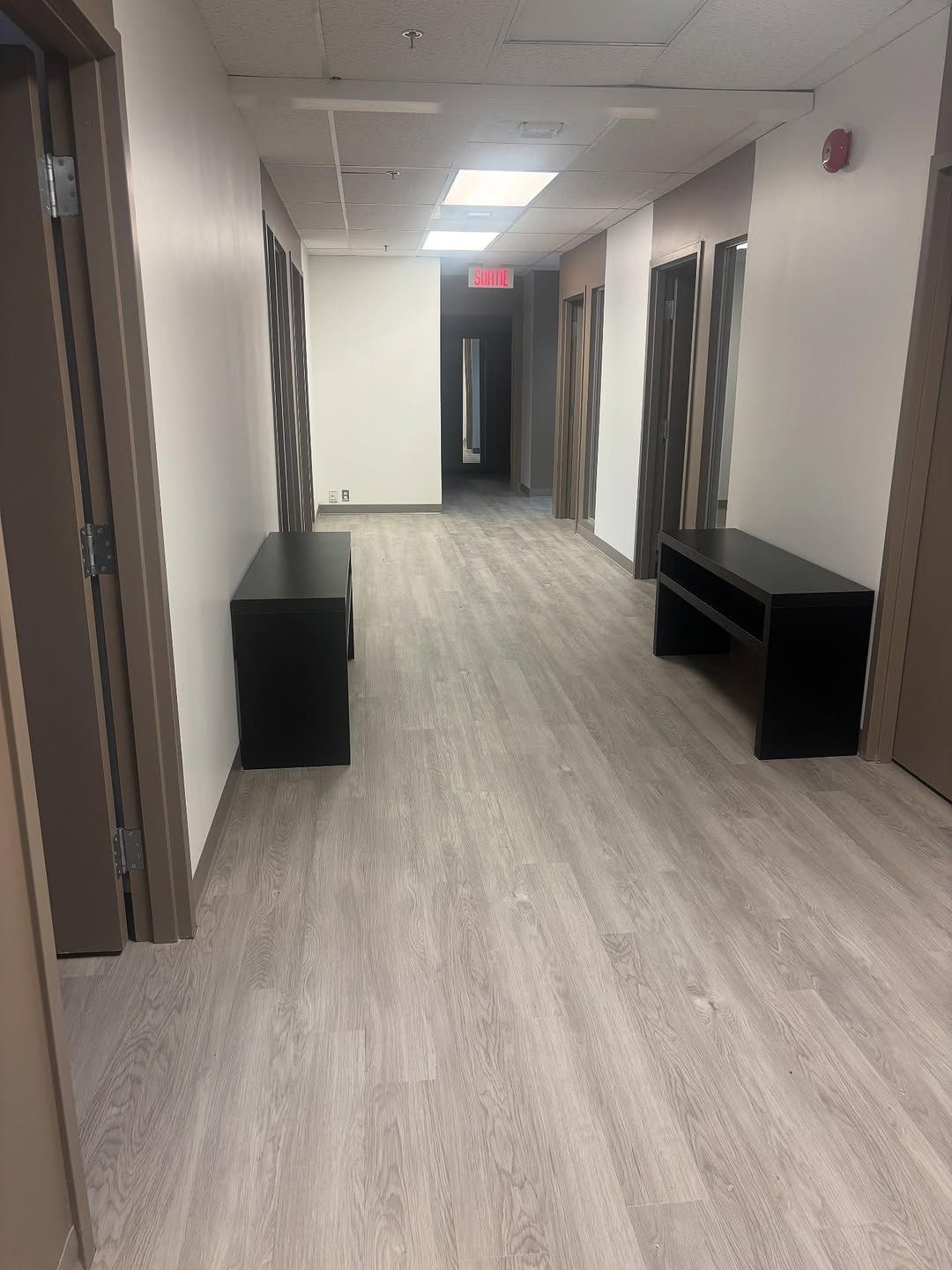 Couloir de bureau vide avec sol gris, portes sombres et bancs noirs éclairés par des néons