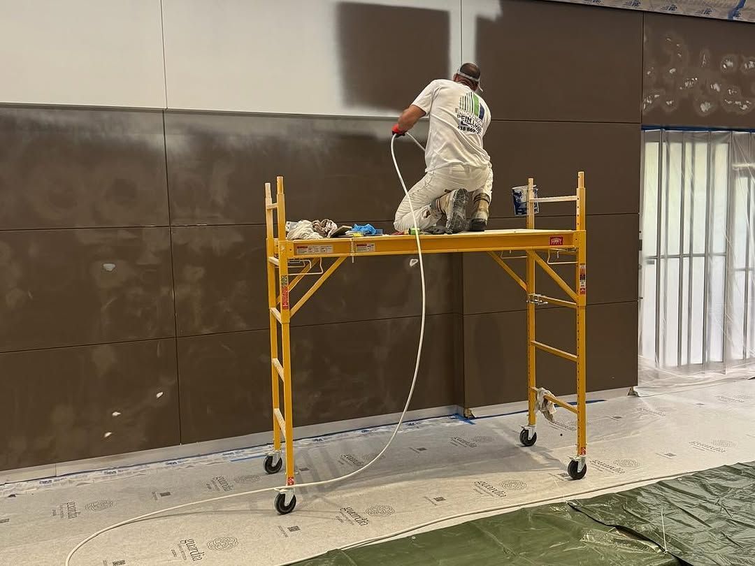 Ouvrier sur un échafaudage jaune en train de peindre un grand mur marron à l'intérieur d'une pièce