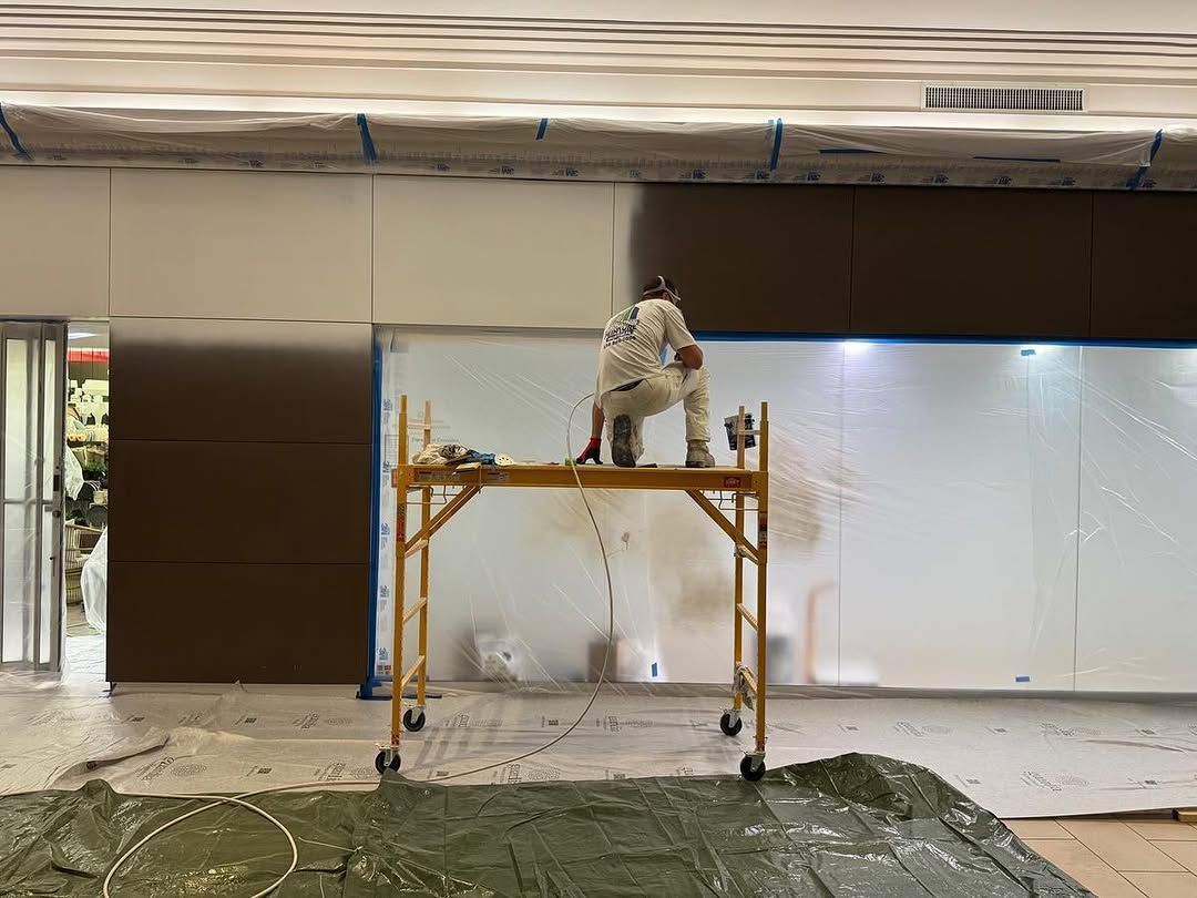 Un ouvrier travaille sur un échafaudage jaune dans une pièce en rénovation, aux murs blancs et recouverte de bâches en plastique.