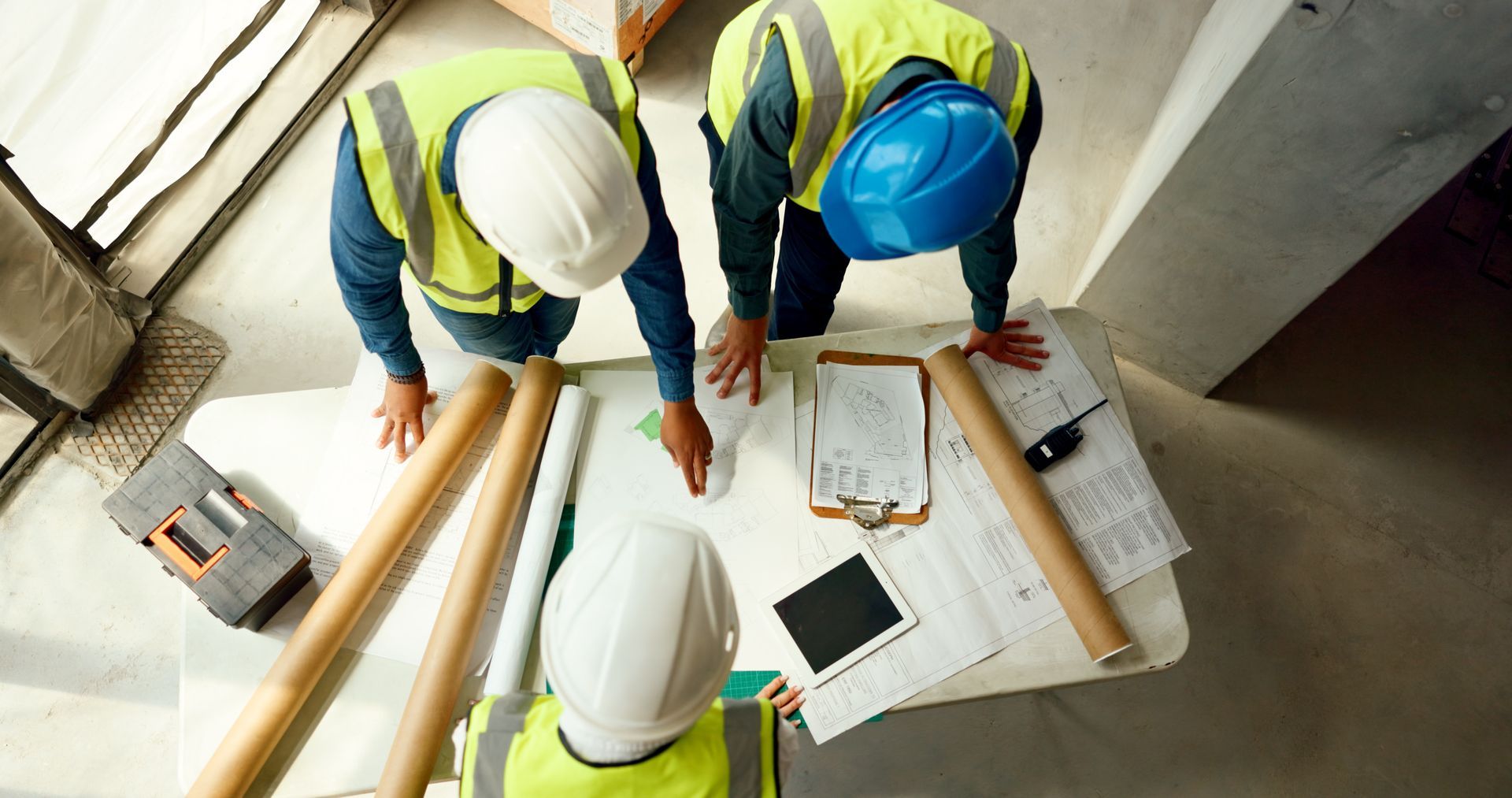 Trois ouvriers du bâtiment, casqués, examinent des plans à une table sur un chantier.